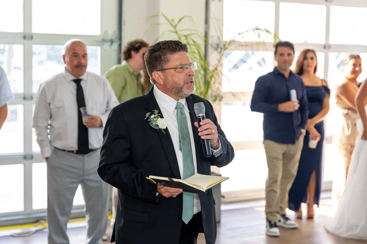 A man in a black suit, glasses, and a light green tie is speaking into a microphone during a wedding reception, holding an open book. Several guests are standing behind him, in a bright room with large windows and plants.
