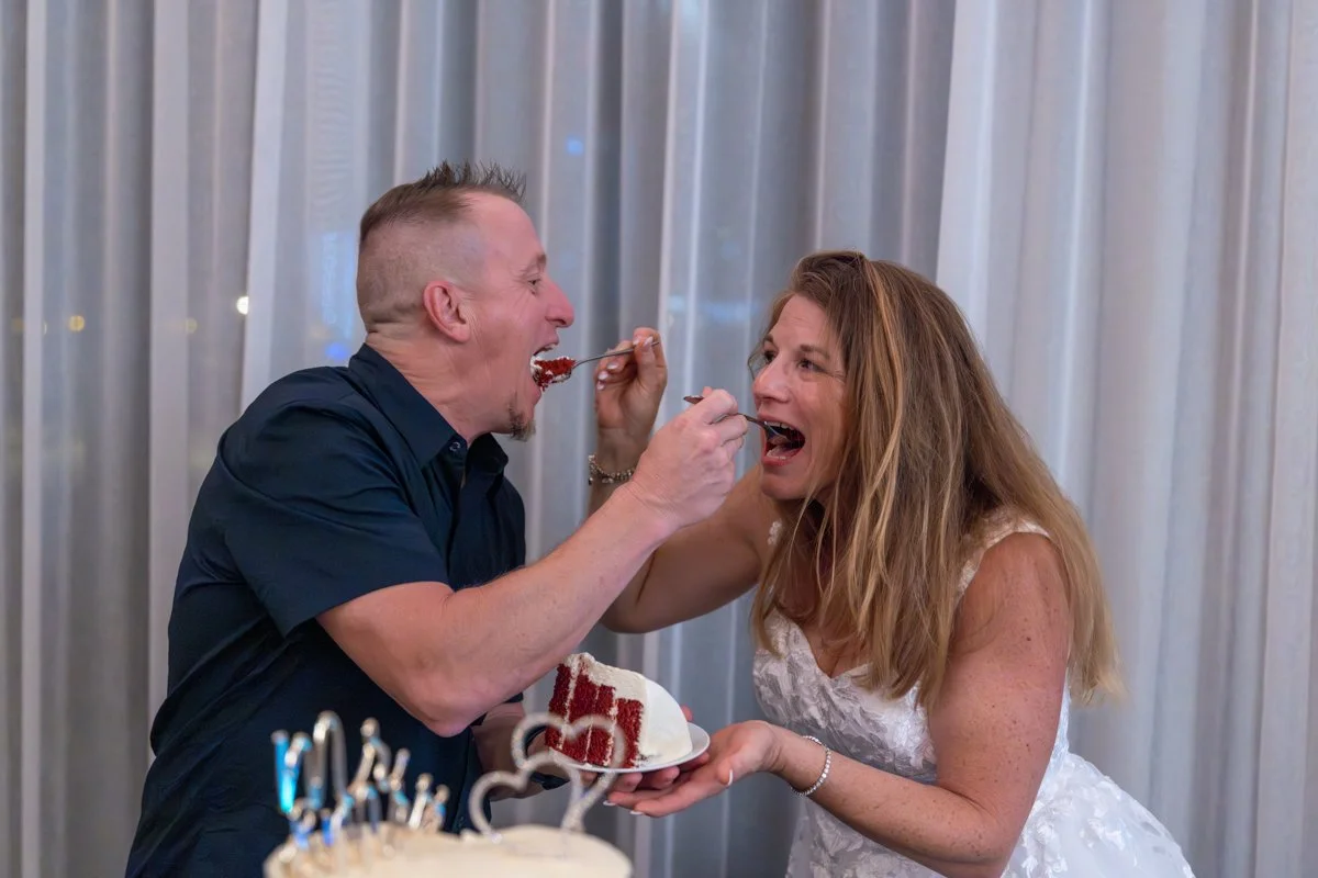 A man and woman celebrate a birthday by feeding each other cake at a party, with a cake topped with birthday candles in the foreground.