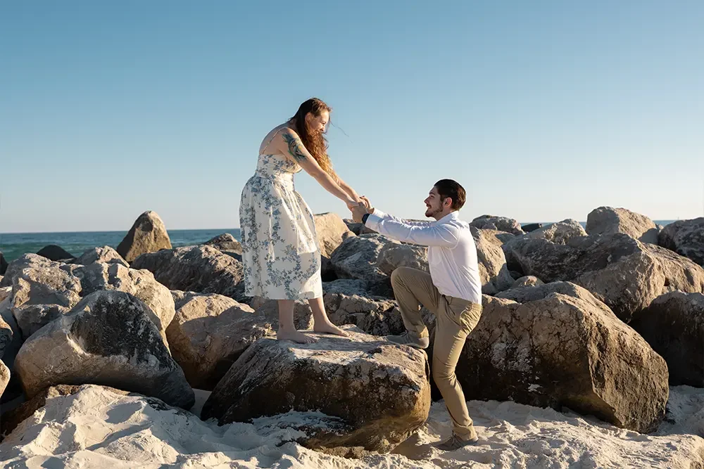 A man on one knee proposing to a woman on a rocky beach during sunset.