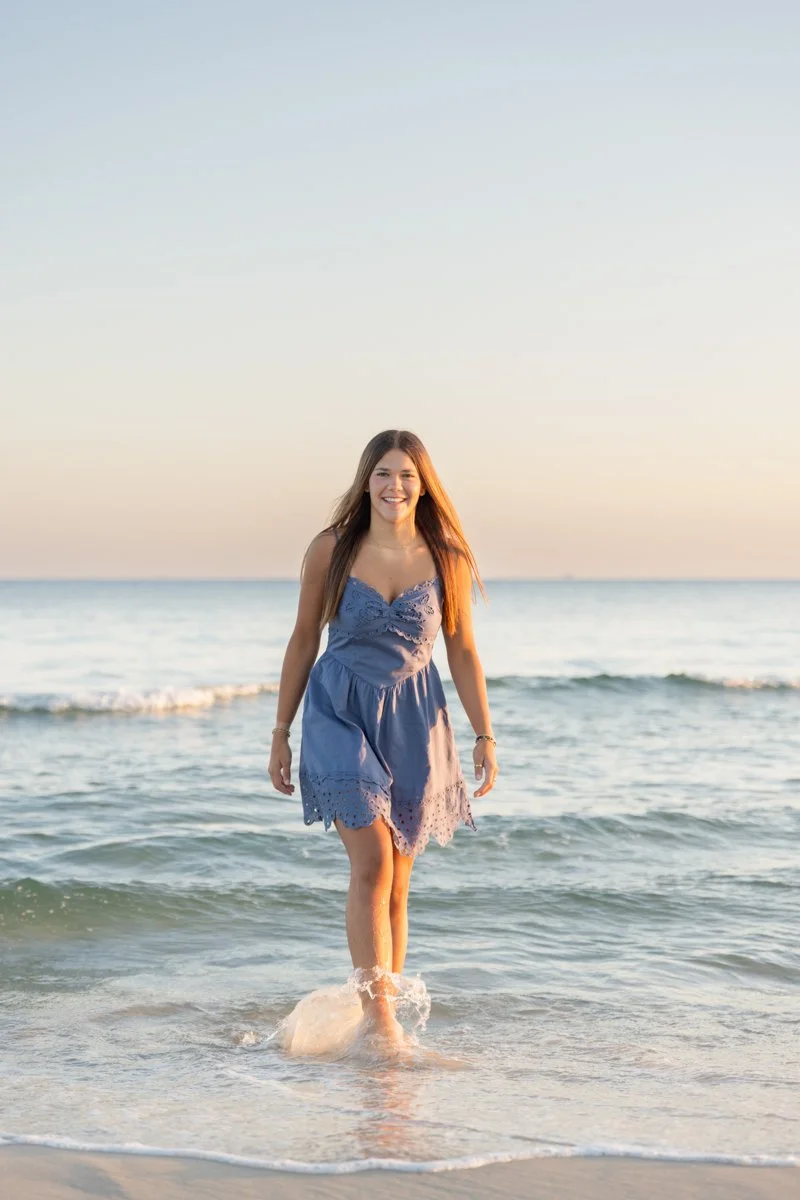 Young woman in a blue dress walking through shallow ocean water at sunset, smiling.