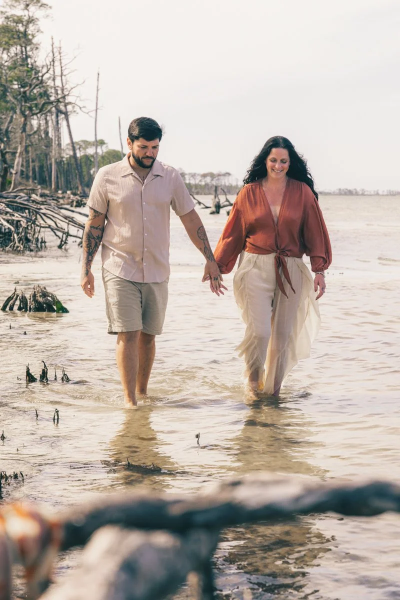 A man and woman holding hands walking in shallow water at a beach with trees and driftwood in the background.