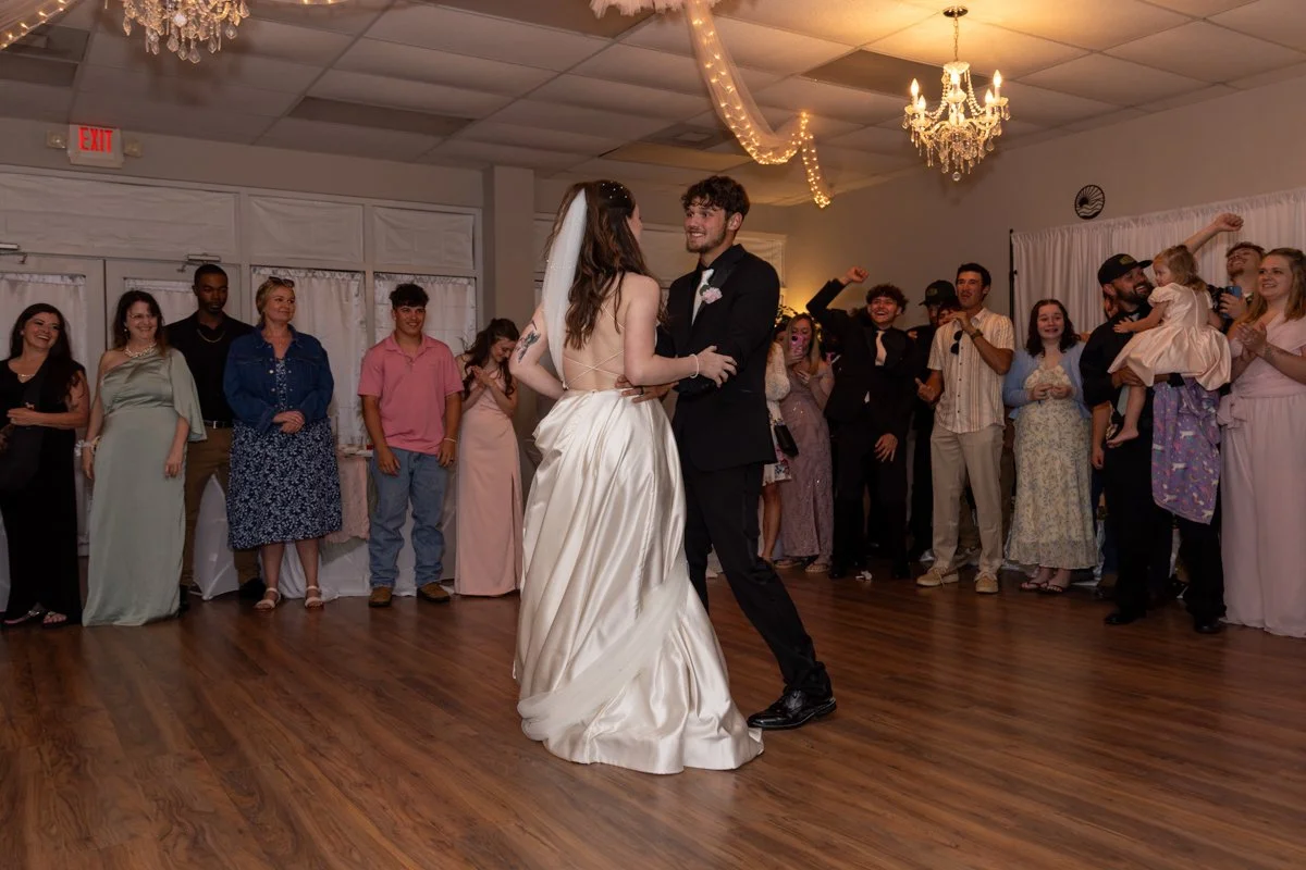 A bride and groom dance together on a wooden floor at their wedding reception, surrounded by guests who are watching and celebrating.