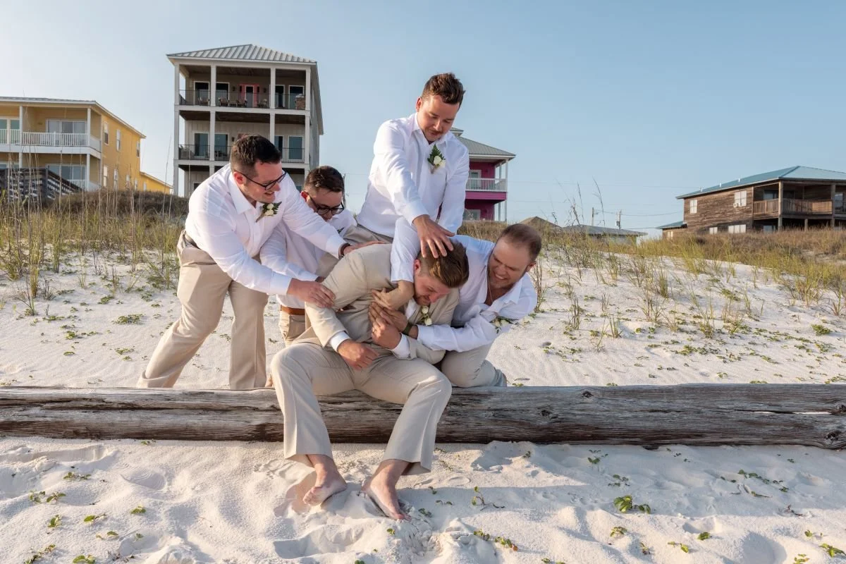 Group of five people in formal attire playfully fighting on a sandy beach with beach houses in the background.