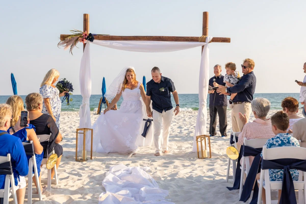 A beach wedding ceremony with a bride and groom walking down the aisle under a wooden arch decorated with fabric and flowers, surrounded by seated guests taking photos and clapping as the ocean is visible in the background.