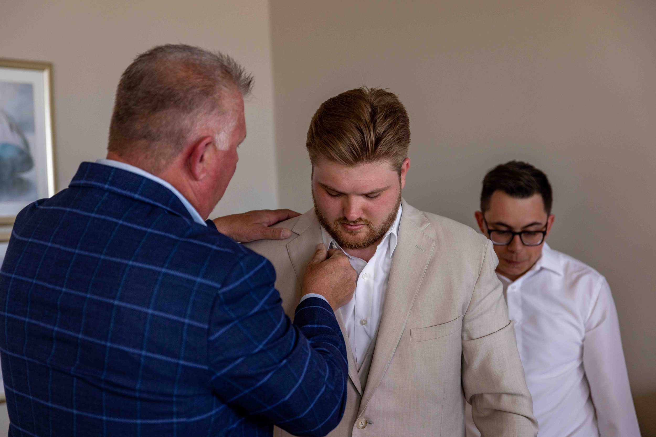 An older man with gray hair and a beard pinning a white boutonniere onto the lapel of a young man with brown hair and a beard, who looks down. Two other men, also dressed formally, are seated in the background.