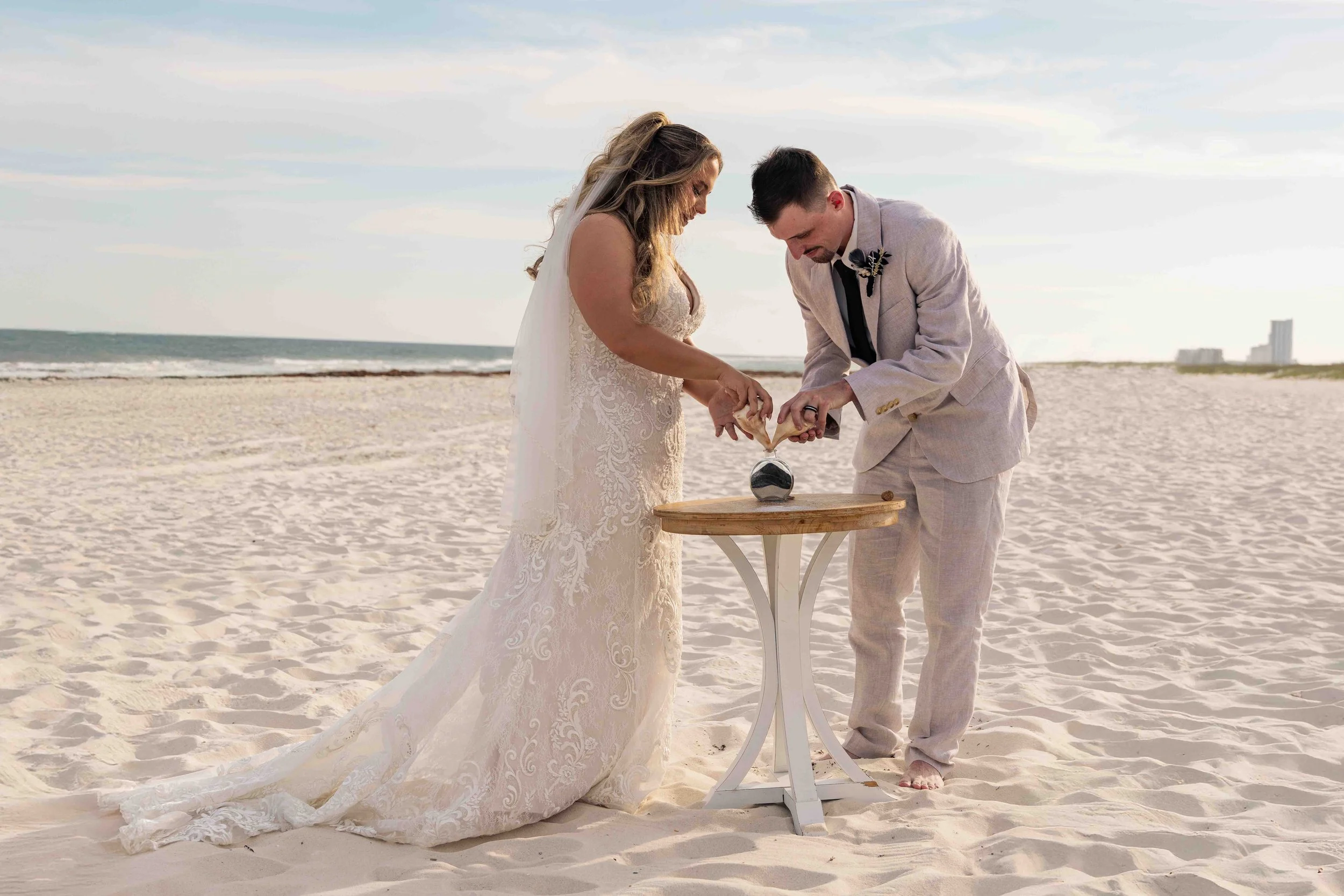 A bride and groom exchanging rings during their beach wedding ceremony at sunset, standing at a small round table on the sand with the ocean in the background.