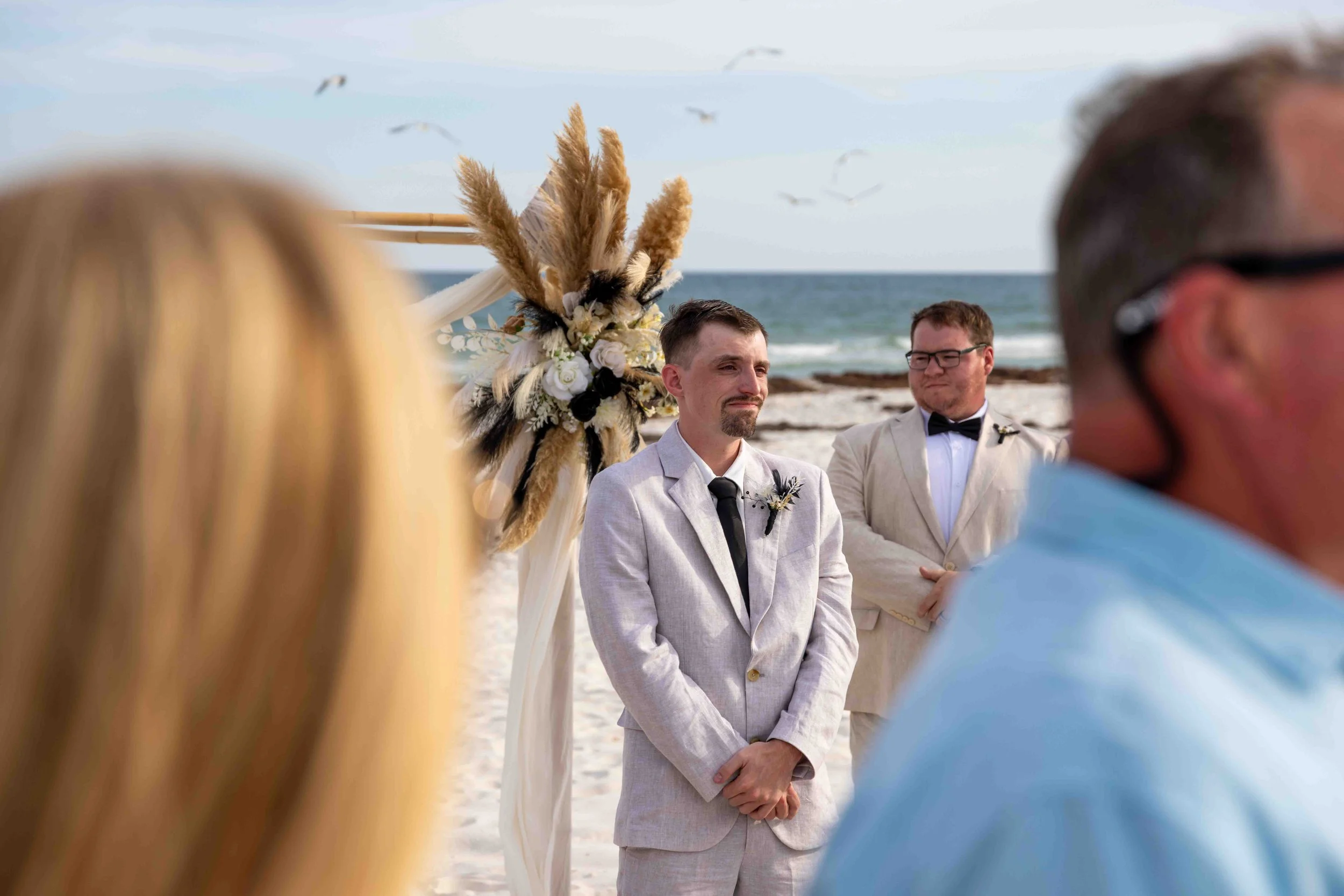 A wedding ceremony on the beach with two men standing in front of an arch decorated with pampas grass, flowers, and raccoon tail accents, with seagulls flying in the background.