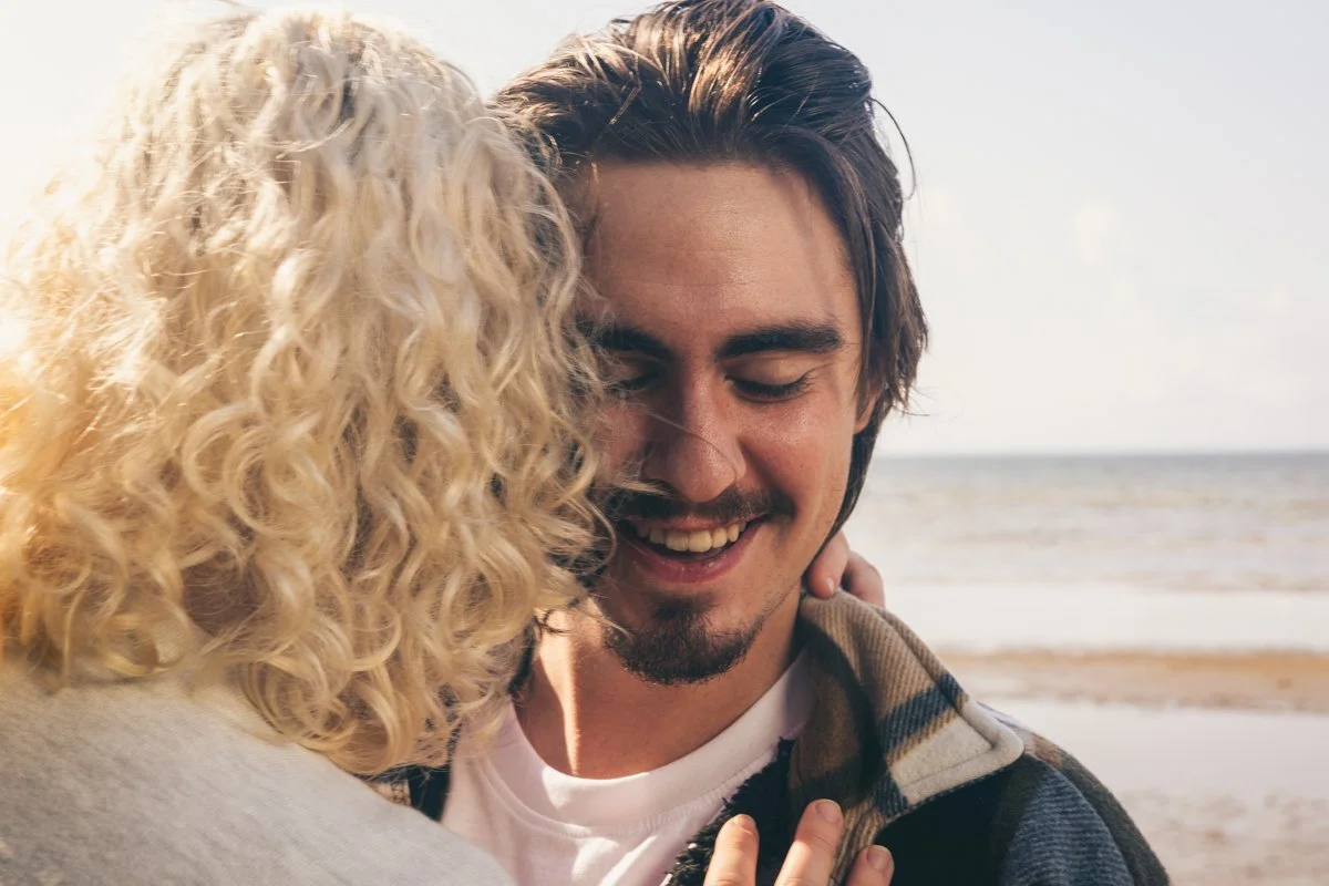 A young man and woman sharing a close, happy moment on the beach, with the woman’s face partially visible and the man smiling with eyes closed.