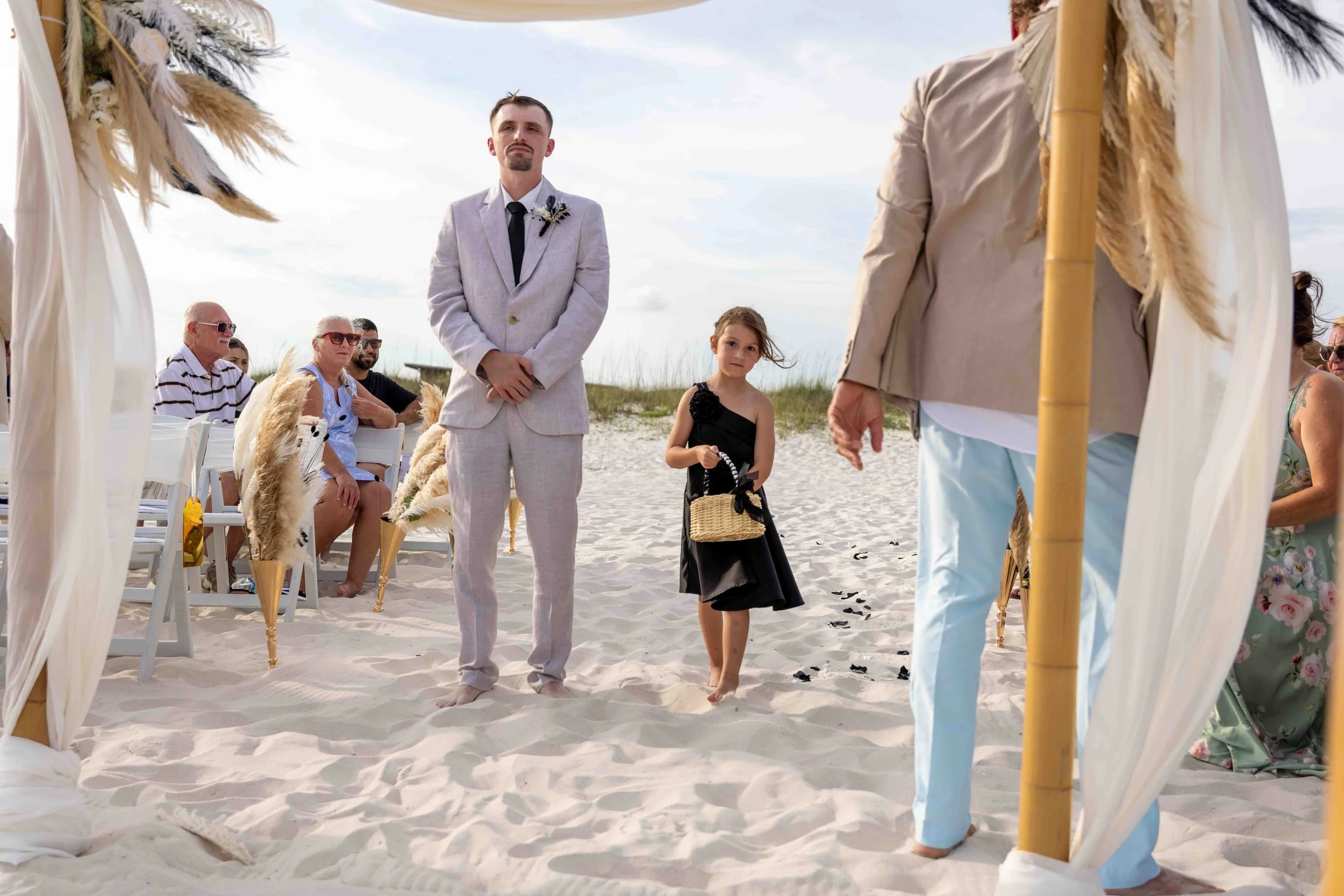 A small wedding ceremony on the beach with a man in a light gray suit standing next to a young girl in a black dress holding a wicker basket. The ceremony is decorated with white fabric, pampas grass, and bamboo poles, with seated guests in the backg