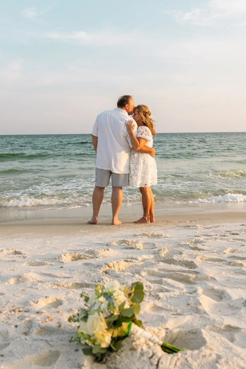 A couple is embracing and kissing on the beach at sunset, with a bouquet of flowers lying on the sand in the foreground.