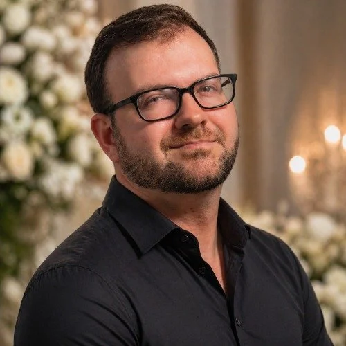 A man with short dark hair, glasses, and a beard, wearing a black shirt, standing indoors with blurred white flowers and warm lighting in the background.