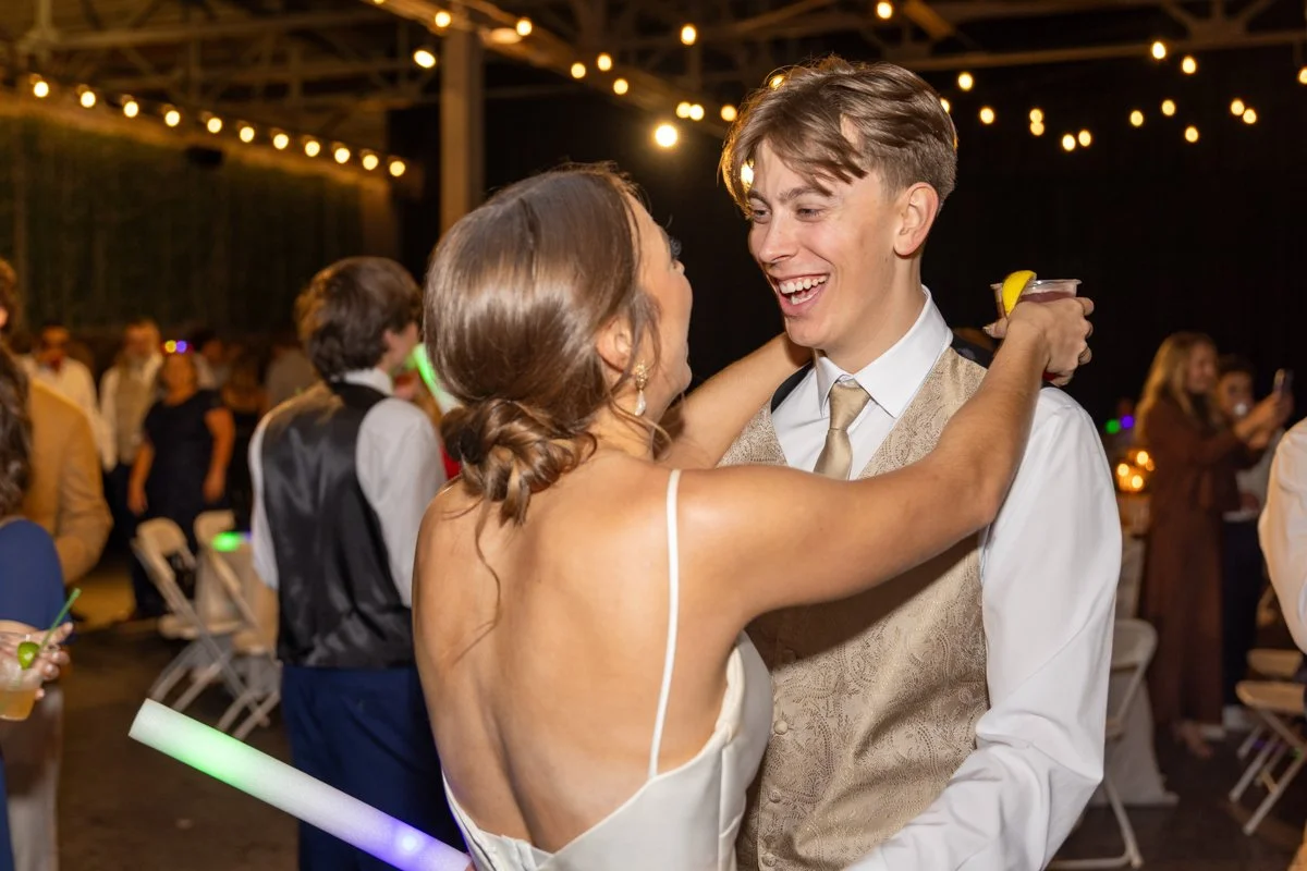 A woman and a young man dancing and smiling at a celebration, with string lights overhead and other guests in the background.
