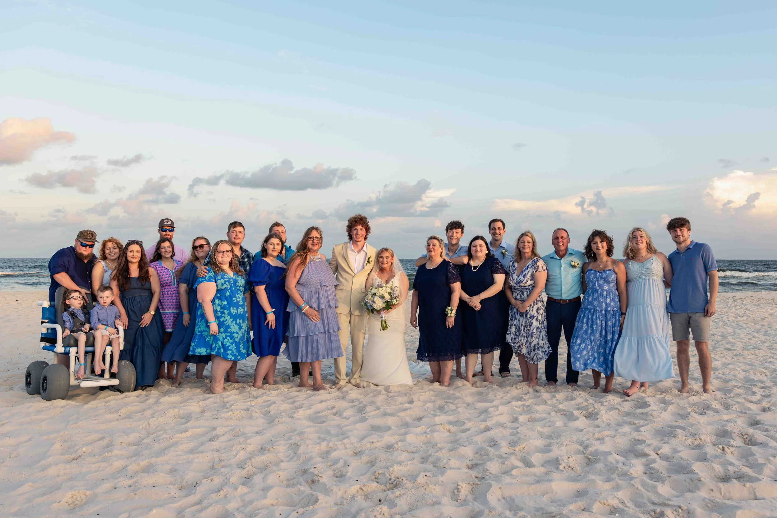 A group of people, including children and adults, gathered on a sandy beach with the ocean and a partly cloudy sky behind them. They are dressed in semi-formal and casual summer clothing, and most are smiling at the camera. The bride in a white dress