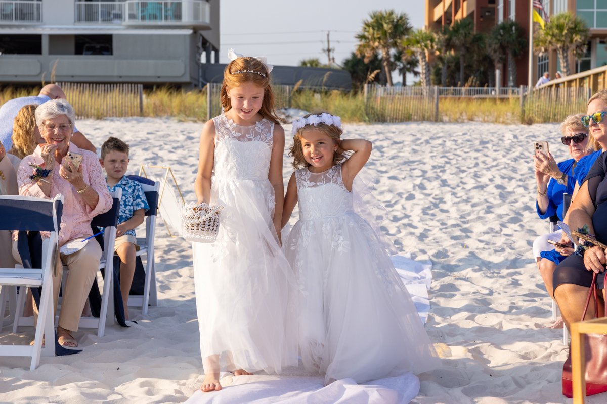 Two young girls in white dresses walking on a beach during a wedding ceremony, with guests seated on either side taking photos and watching.