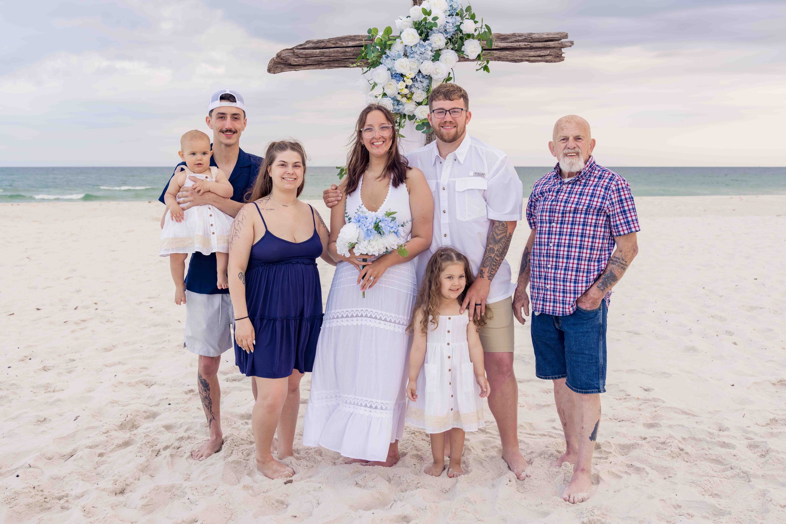 A group of seven people, including children, adults, and an elderly man, standing on a sandy beach in front of a floral wedding arch with the ocean in the background.
