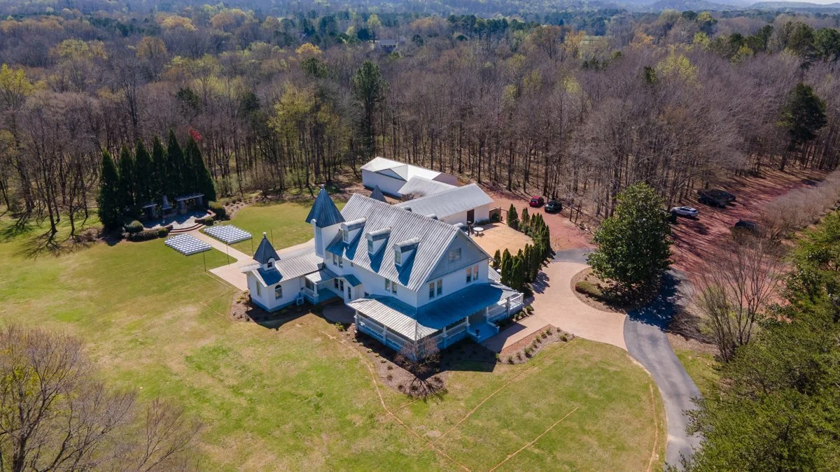 A large white house with multiple steeply pitch roofs and a tower, situated in a spacious rural landscape surrounded by trees, with a circular driveway, a lawn, and several outbuildings or garages. There are parked cars near the outbuildings and a wo