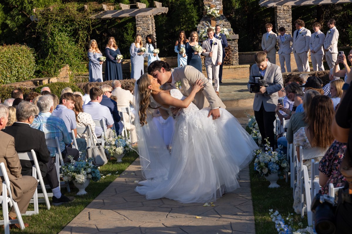 A bride and groom sharing a kiss at their outdoor wedding ceremony, with guests seated on either side and wedding party members standing in the background.