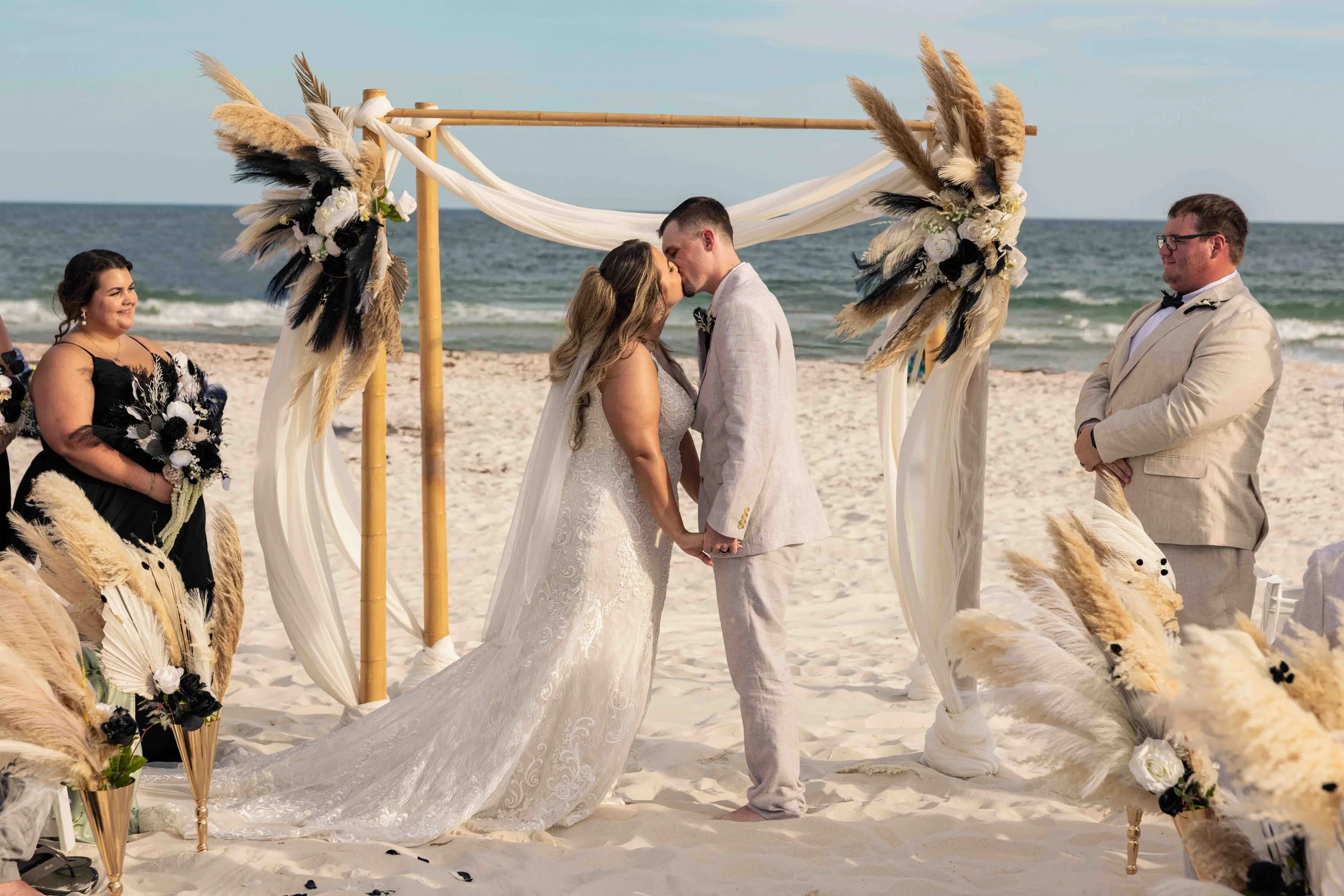 A couple is kissing during their beach wedding ceremony with the ocean in the background. The bride is in a white lace wedding dress, and the groom is in a light-colored suit. There are wedding officiants and a decorative arch with flowers and feathe