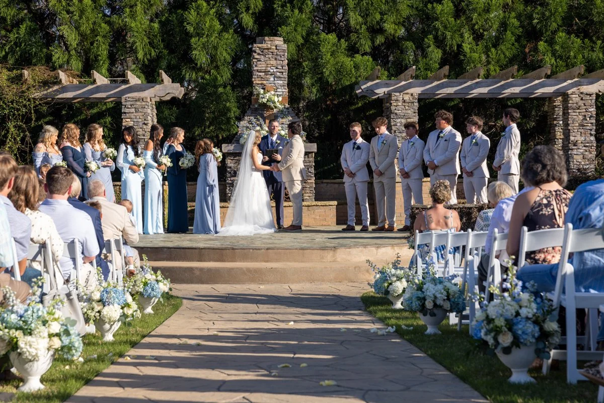 A wedding ceremony outdoors with the bride and groom exchanging vows, surrounded by bridesmaids and groomsmen, with guests seated on white chairs decorated with white and blue flowers.