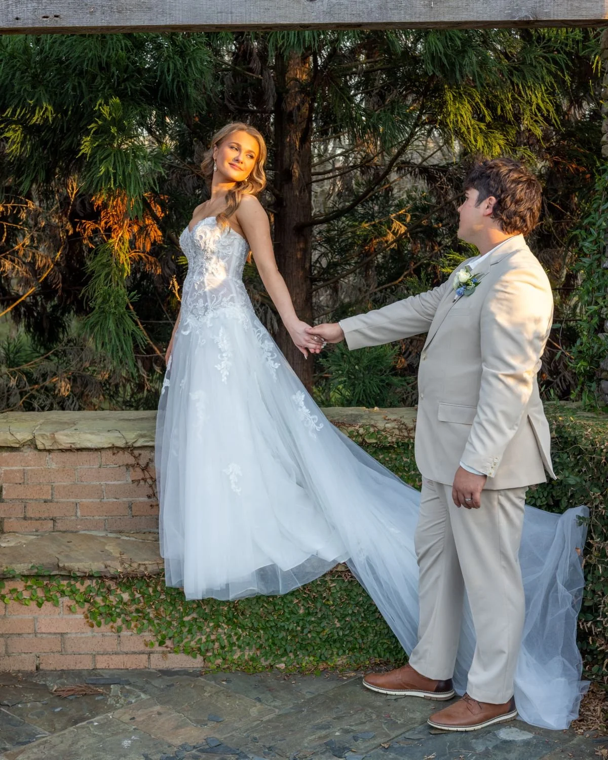 A bride and groom holding hands outdoors, with the bride wearing a white wedding dress and the groom in a light tan suit, standing in front of green trees and a brick wall.