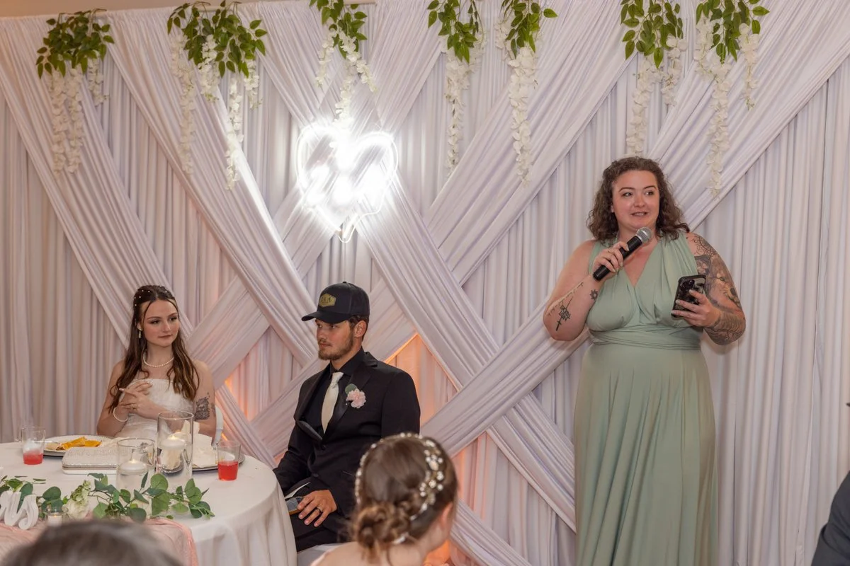 A woman with tattoos in a light green dress is giving a speech at a wedding reception, holding a microphone in one hand and a phone in the other. A bride wearing a white dress and veil and a groom in a suit and cap are sitting at a table decorated wi