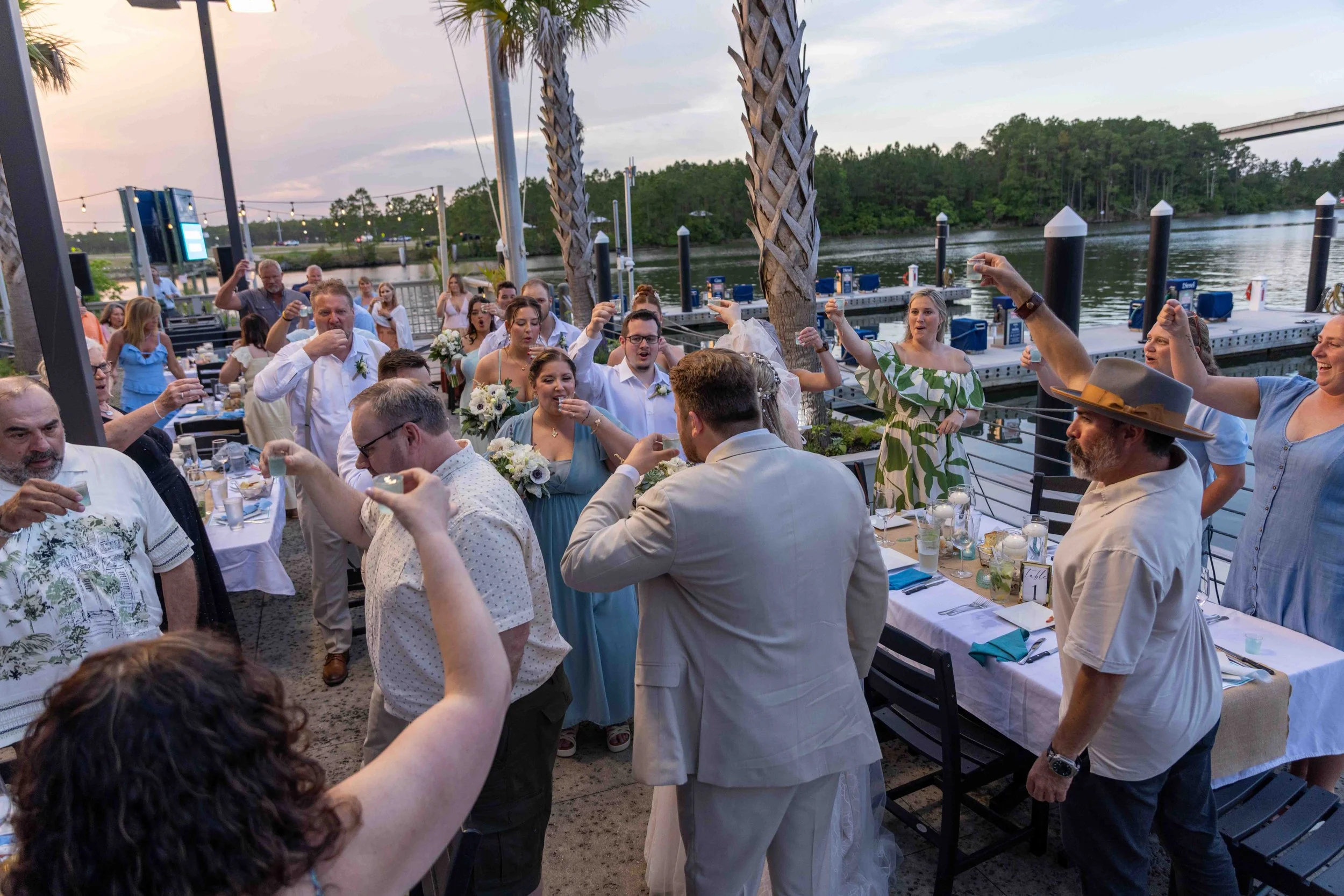 A group of people celebrating outdoors near a river, with some raising glasses in a toast, during what appears to be a wedding reception or party, at sunset.
