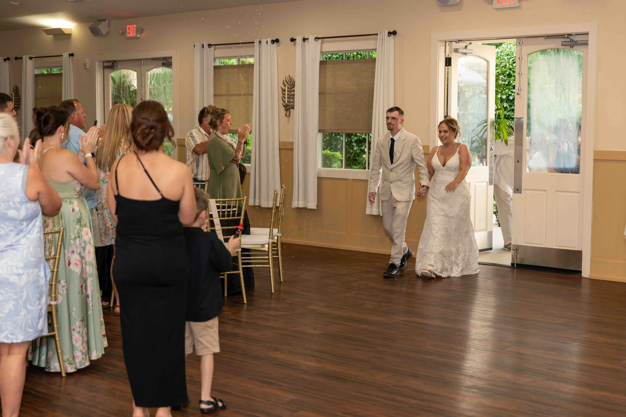A bride and groom walking into a room full of guests at a wedding reception, holding hands and smiling.