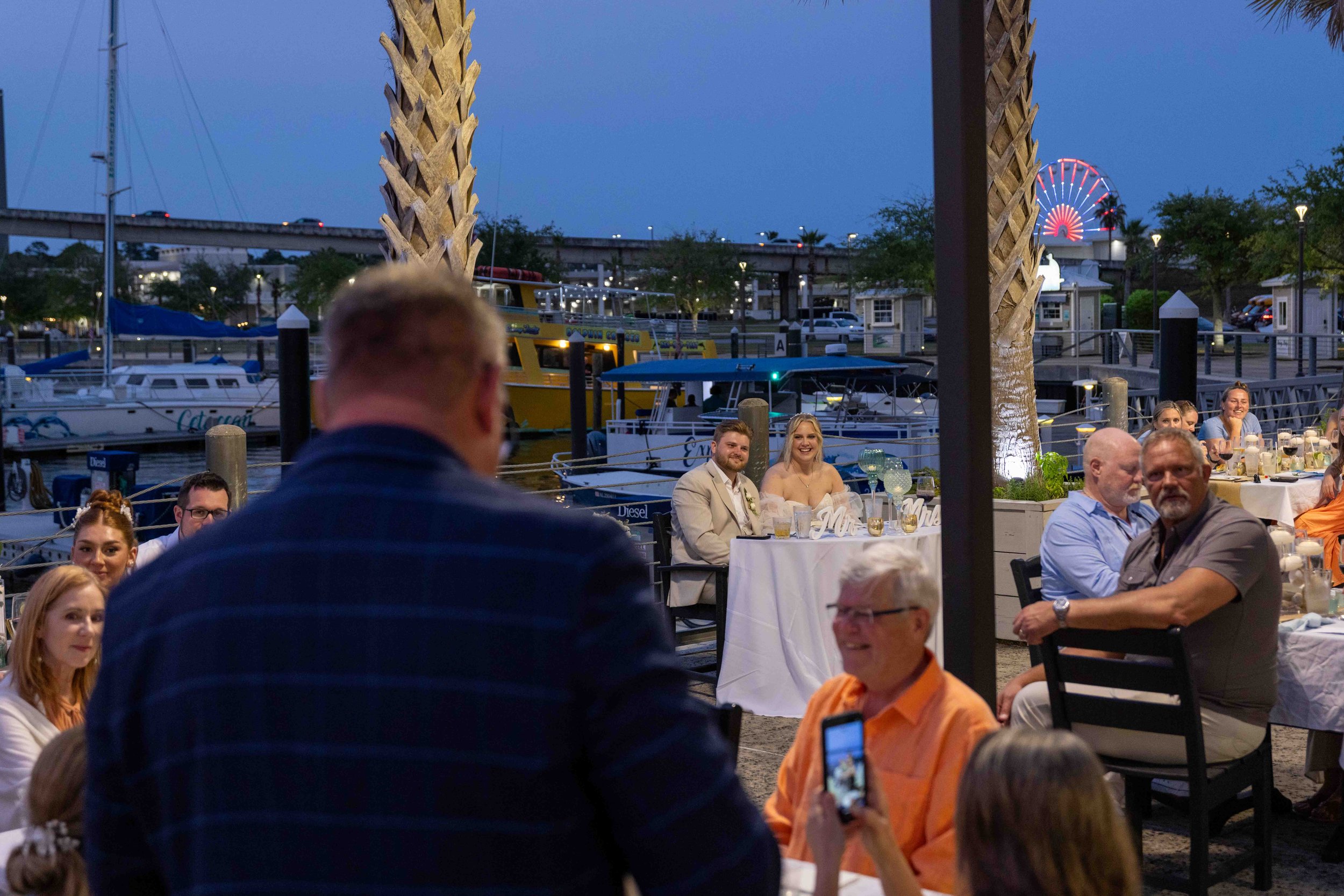 People gathered at an outdoor waterfront dinner event during evening with boats docked nearby and a lit Ferris wheel in the background.