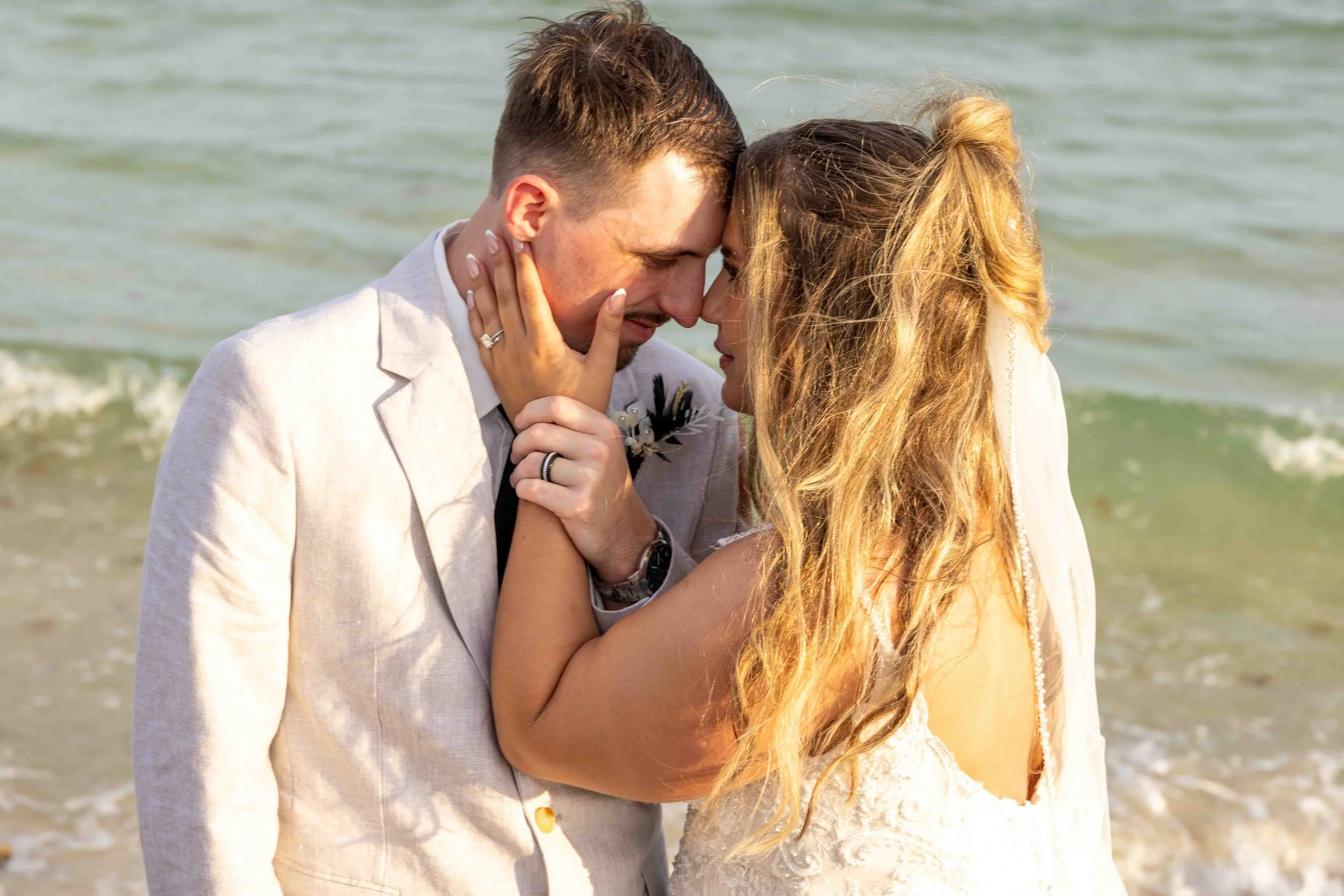 A man and woman share an intimate moment on the beach, touching foreheads and holding each other's faces, with the ocean in the background.