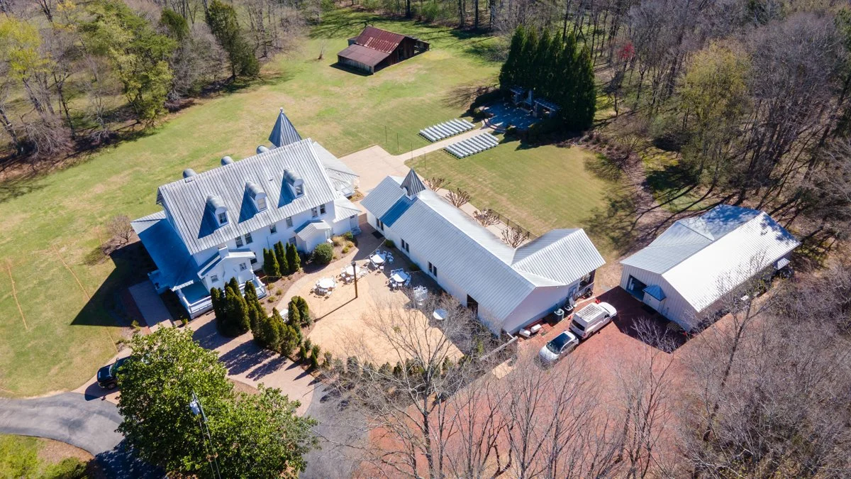A large white mansion with a metal roof, surrounded by a landscaped yard with trees and a patio area with outdoor furniture. There are multiple outbuildings and a driveway with parked cars. The property is bordered by a wooded area.
