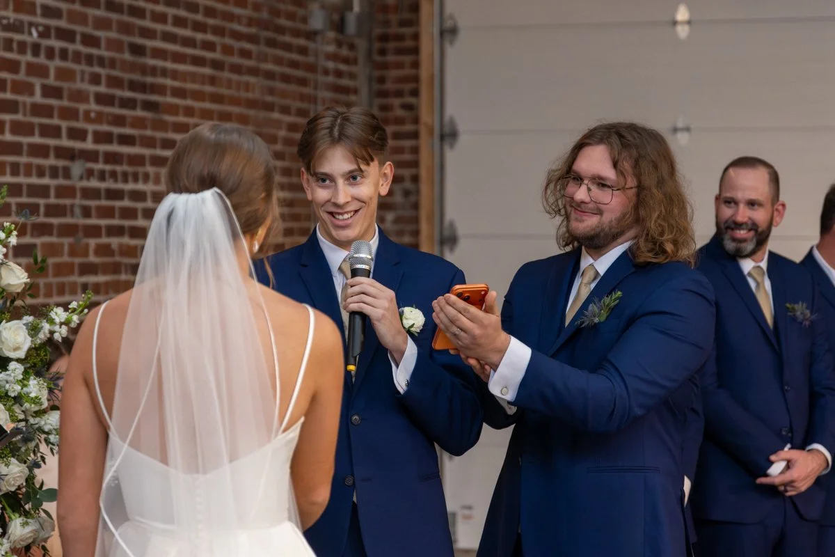 A bride in a white wedding dress with a veil stands facing a groom in a blue suit during a wedding ceremony. A man with glasses in a matching blue suit holds a microphone for the groom, while another man in a similar suit looks on and holds a phone. 
