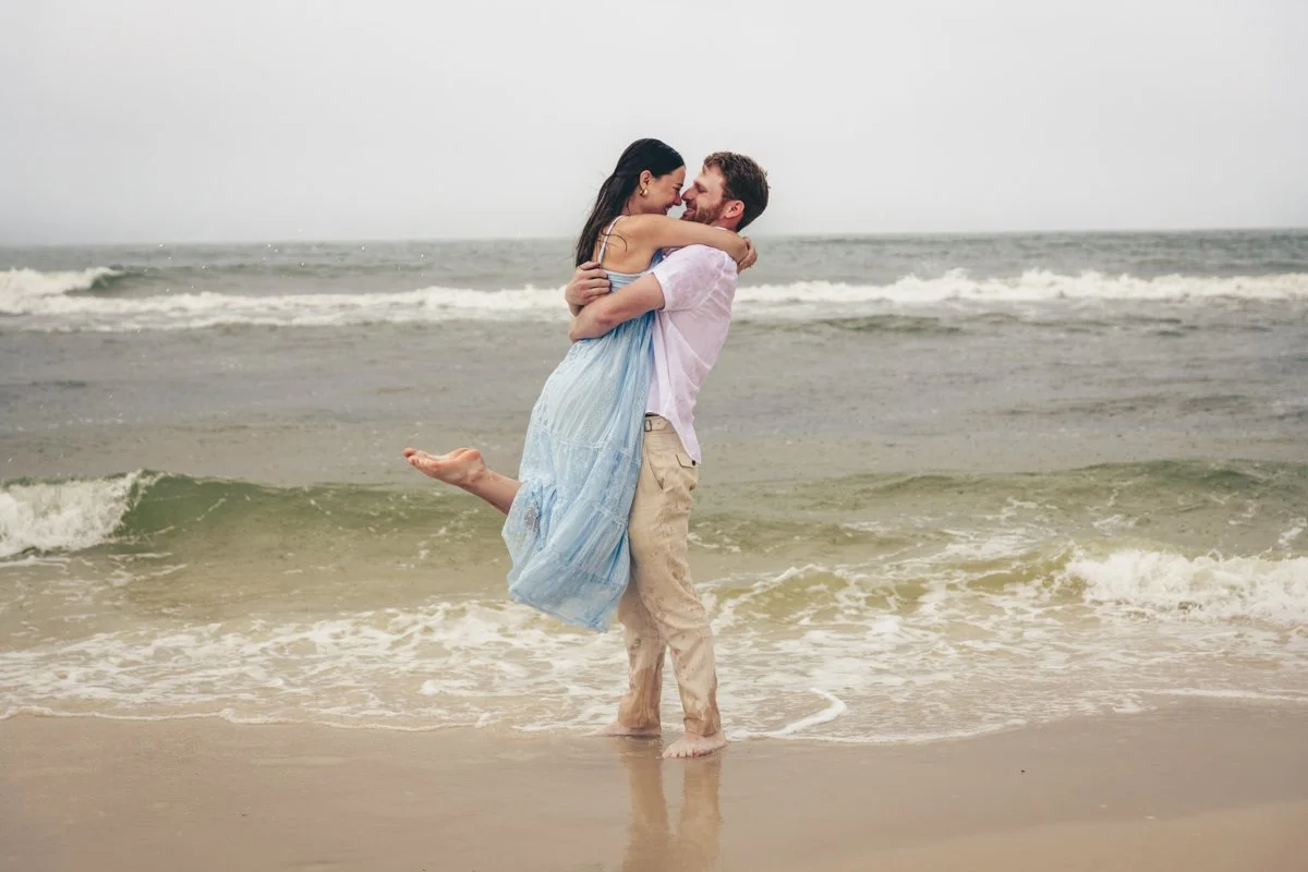 A man lifting a woman in a blue dress on the beach with ocean waves in the background, both smiling and looking at each other.