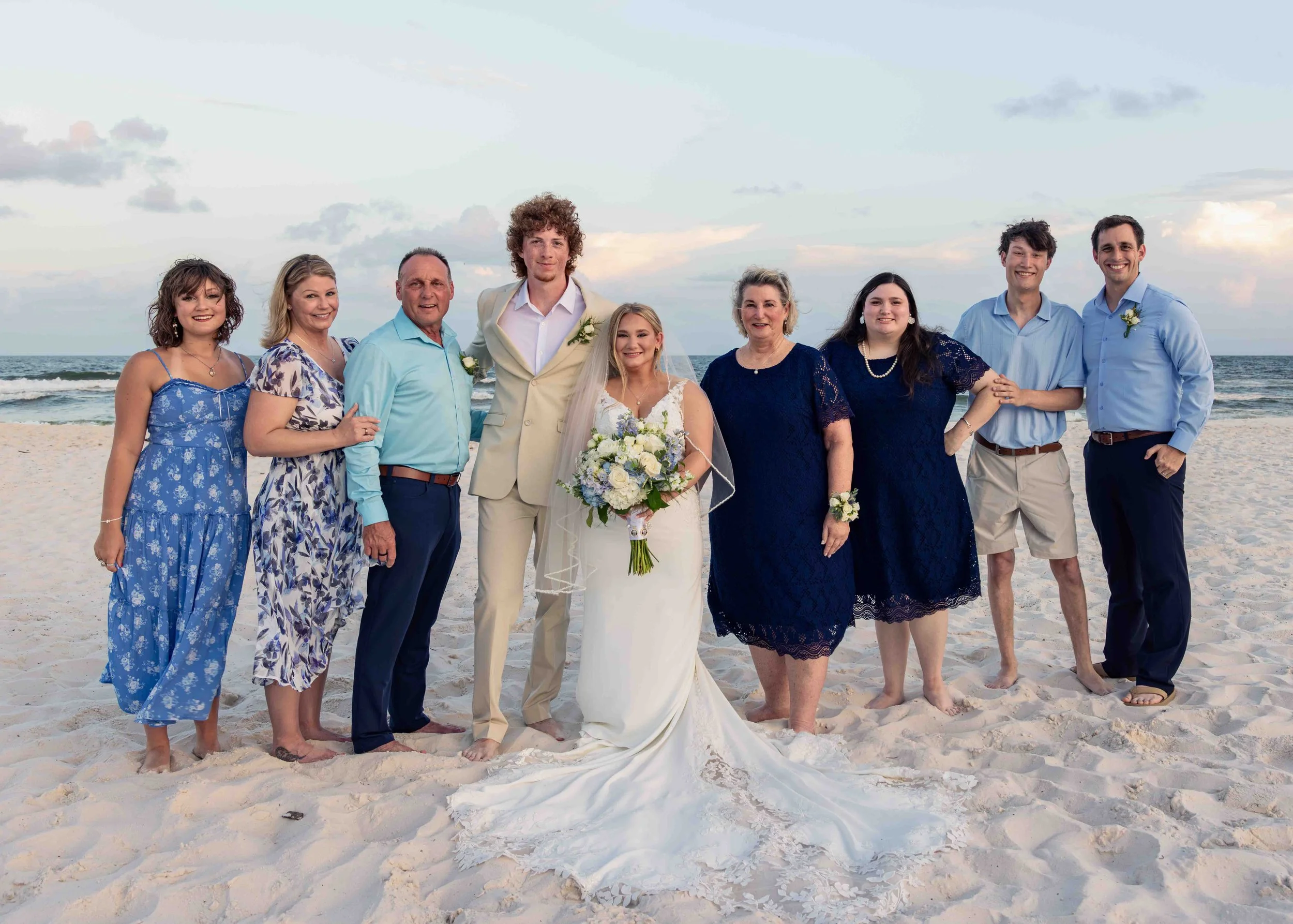 Group of people at a beach wedding, standing on sand with the ocean and sky in the background.