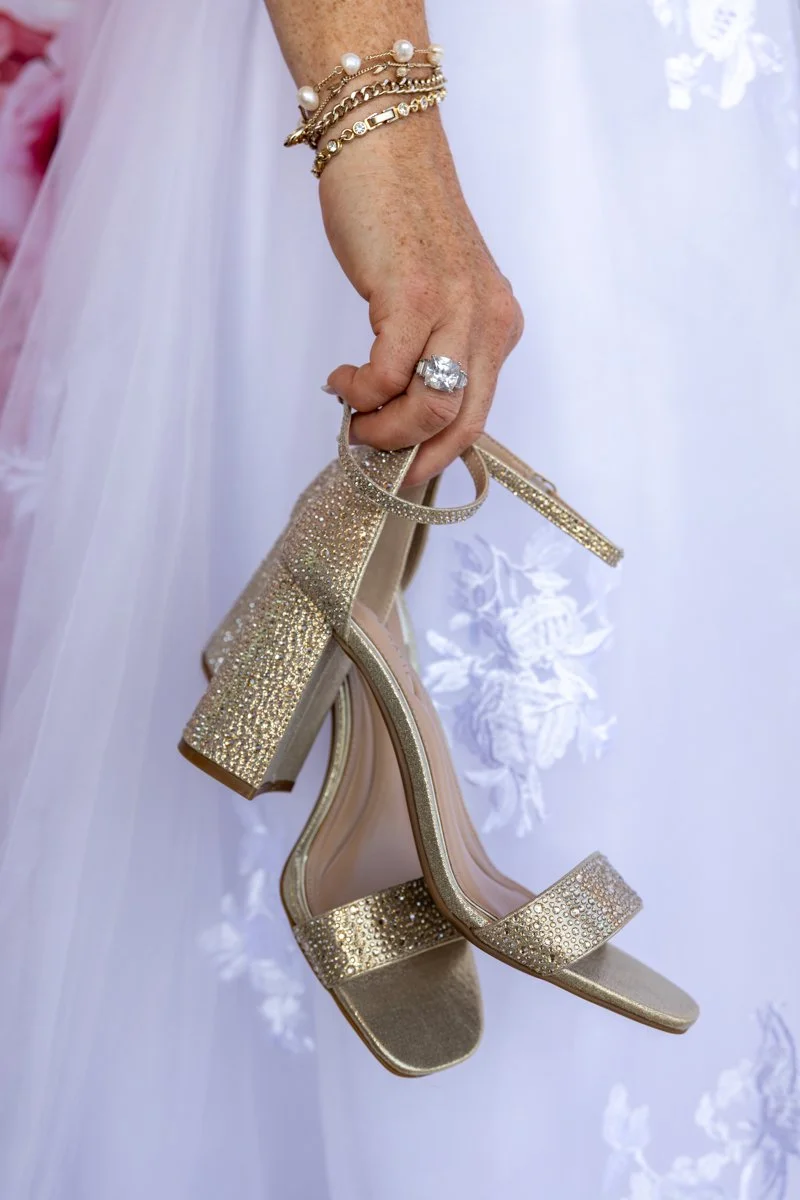 A hand holding a pair of gold high-heeled sandals with sparkling details, wearing a large diamond ring and gold bracelets, with a white floral-patterned dress in the background.