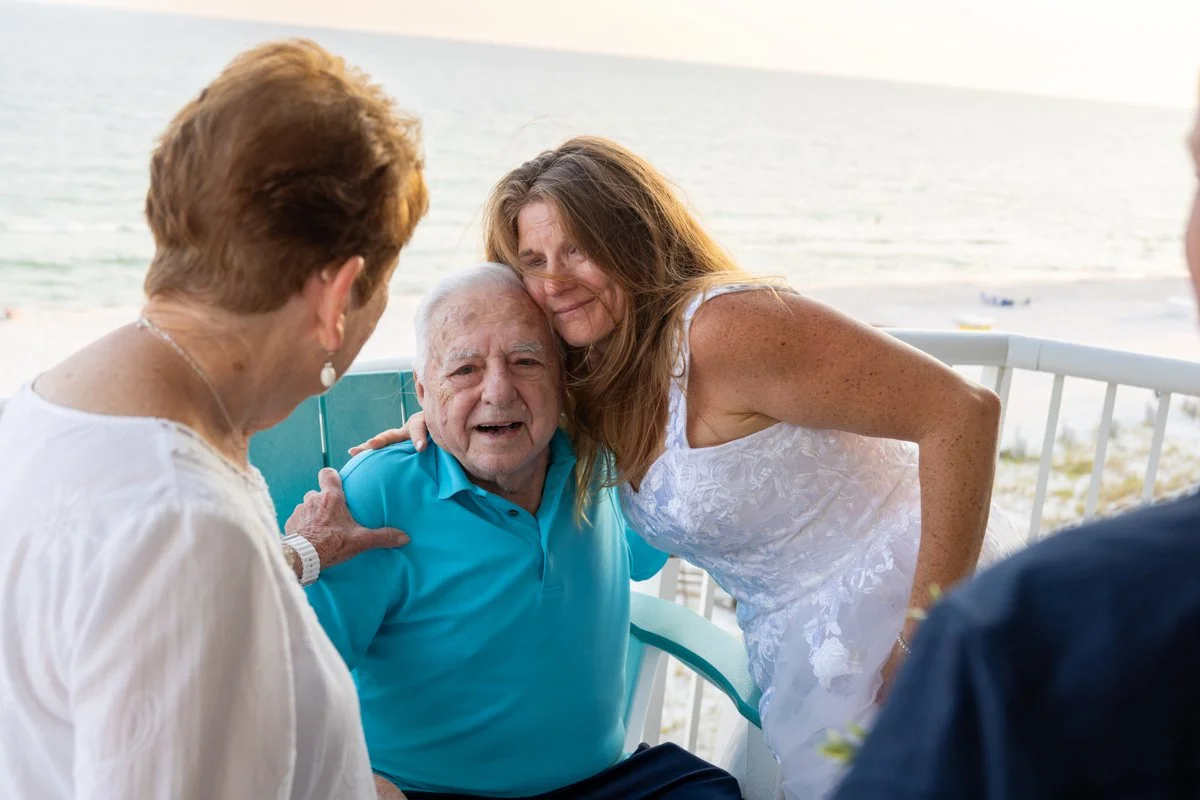 Family gathered on a beachside porch, smiling and hugging an elderly man in a blue polo shirt.