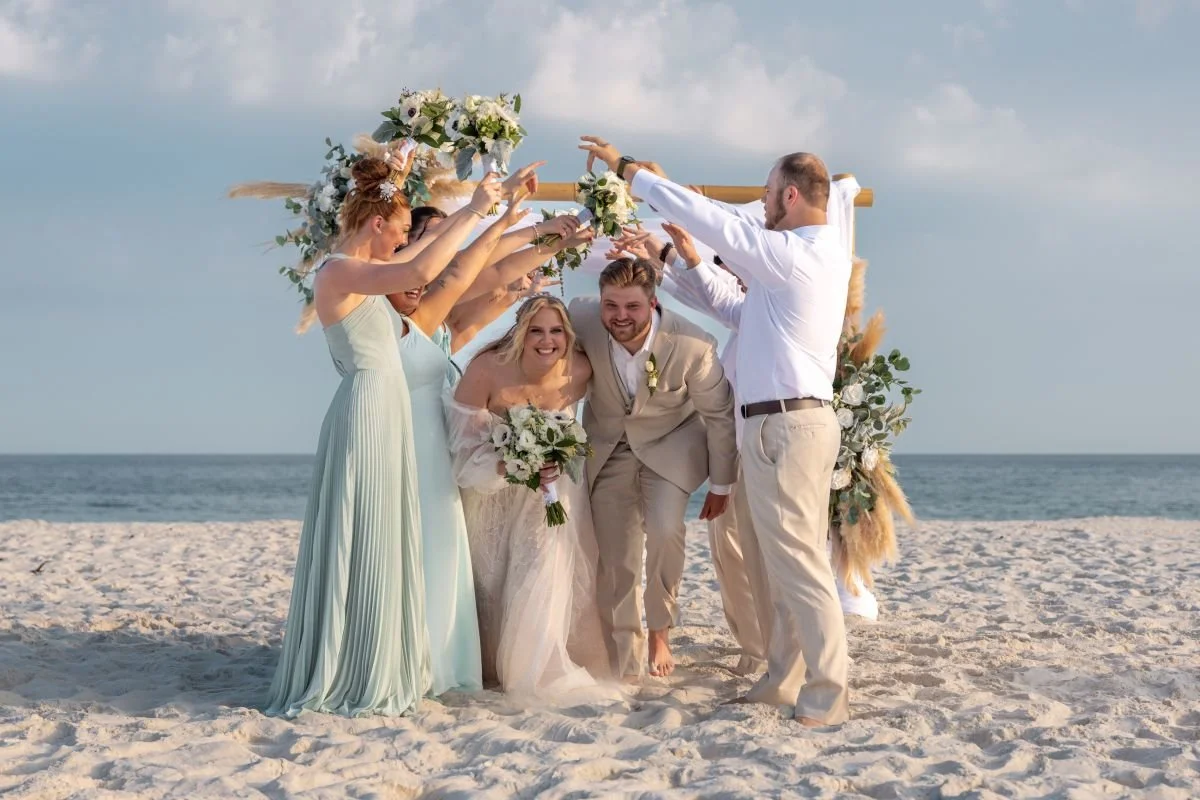 A wedding party on a beach with a floral arch, including the bride in a white wedding dress holding a bouquet, the groom in a beige suit, and four bridesmaids in pastel dresses and a groomsman in a white shirt and beige pants, all smiling and celebra