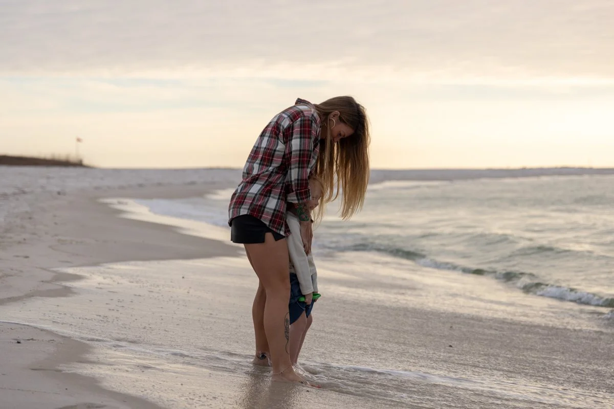 A woman and a child standing together on a beach near the water, with the woman helping the child shoes off.
