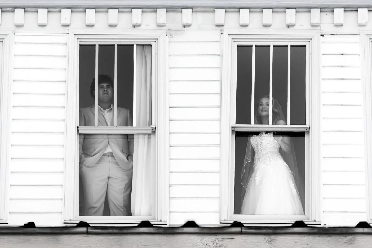 A bride and groom looking out of windows of a house, seen from outside, black and white photograph.