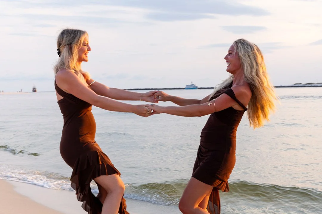 Two women in black dresses holding hands and dancing on the beach at sunset, smiling and facing each other with water and boats in the background.