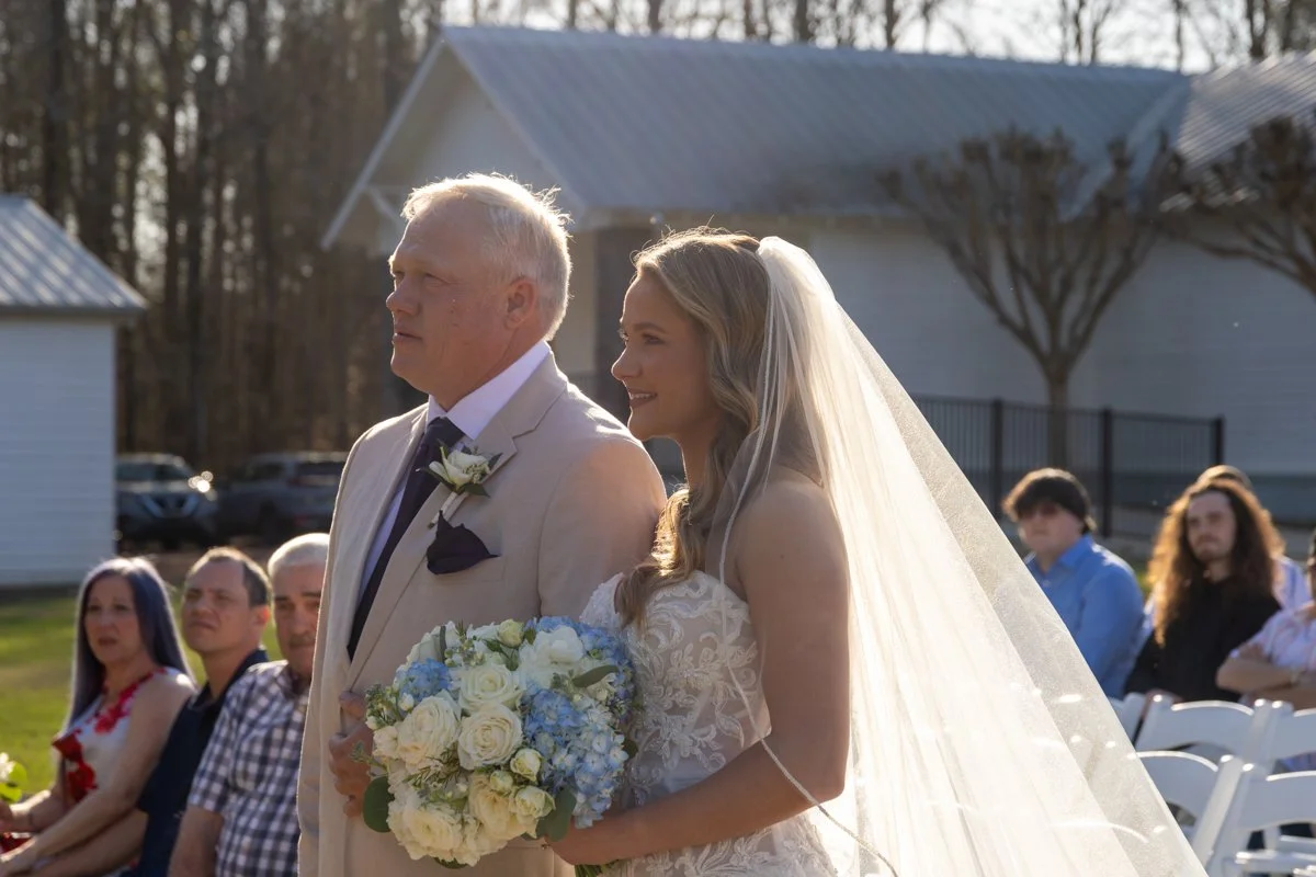 A bride walking down the aisle with her father at an outdoor wedding ceremony. The bride holds a bouquet of white and blue flowers, and the father wears a beige suit with a boutonniere. Guests are seated in the background, and a white building is vis