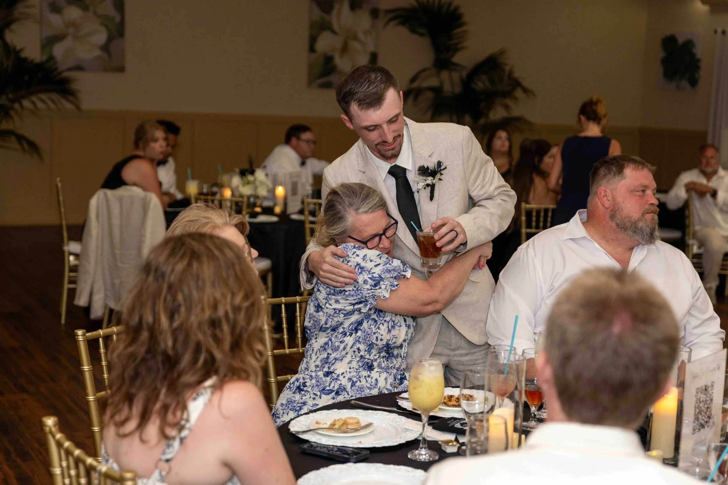 A wedding reception scene with guests sitting at decorated tables, some with drinks and desserts, and a man in a beige suit and black tie giving a hug to an older woman with glasses, a floral dress, and gray hair, surrounded by other guests.