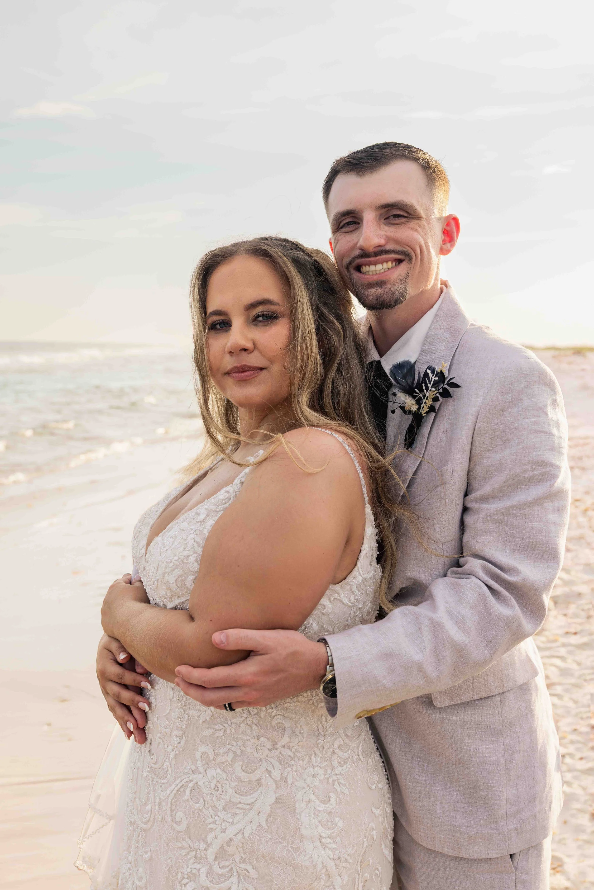 A smiling couple, woman in a white lace wedding dress and man in a light gray suit with boutonniere, standing on a beach with the ocean and cloudy sky in the background.