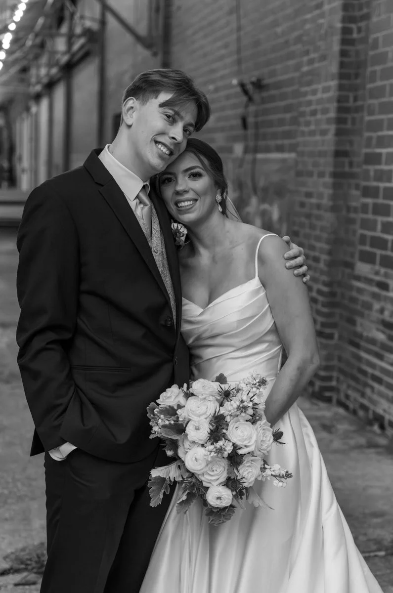 A black and white photo of a bride and groom smiling and hugging outdoors, with a brick wall in the background. The bride is holding a bouquet of flowers.