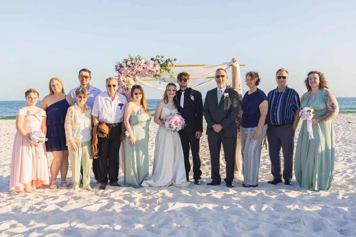 Group of people dressed in formal attire posing on a beach in front of a wedding arch with pink flowers and white drapery during a wedding ceremony.