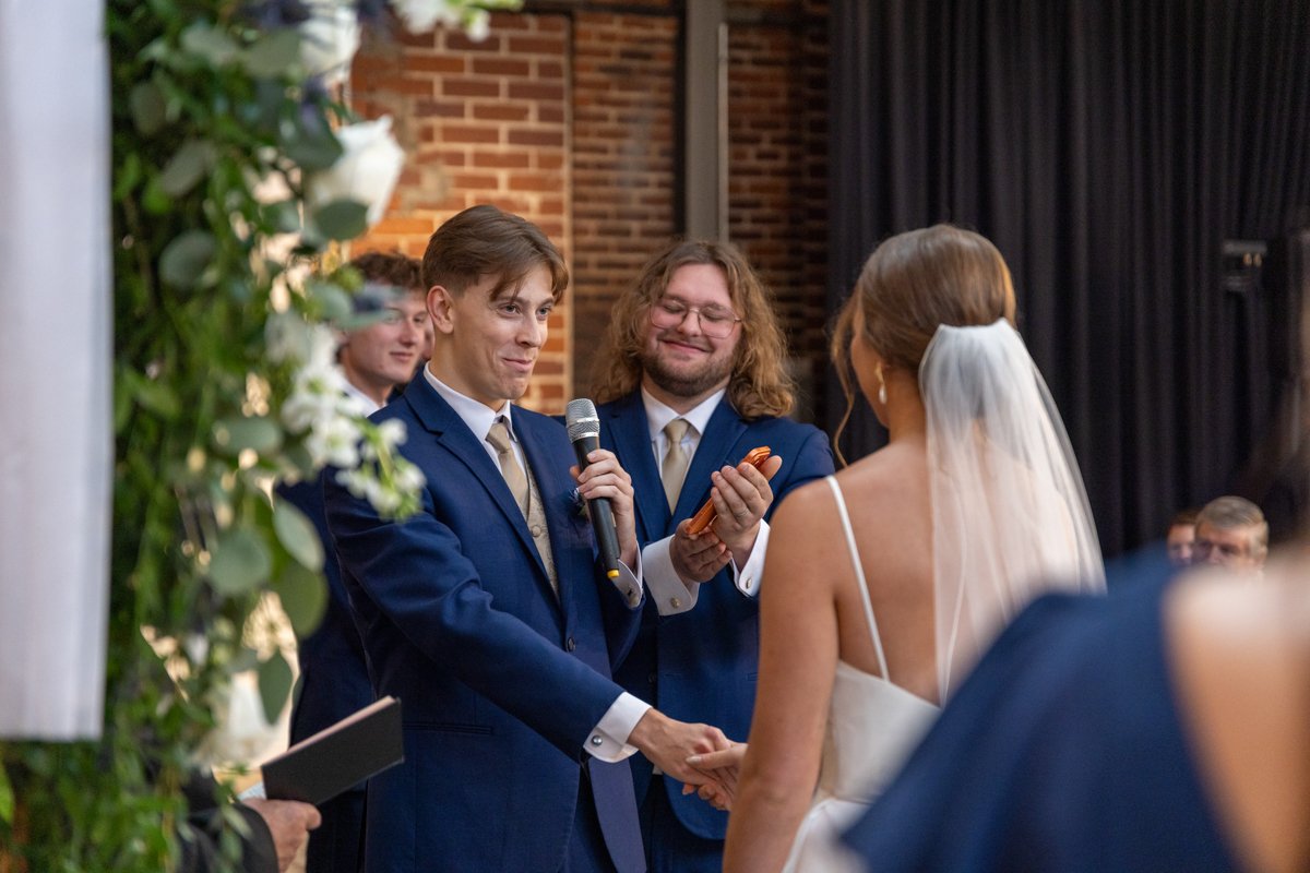 A wedding ceremony with a groom in a blue suit holding the bride's hands while a man with long hair and glasses reads from a paper, all smiling. The bride is wearing a white dress with spaghetti straps and a veil.