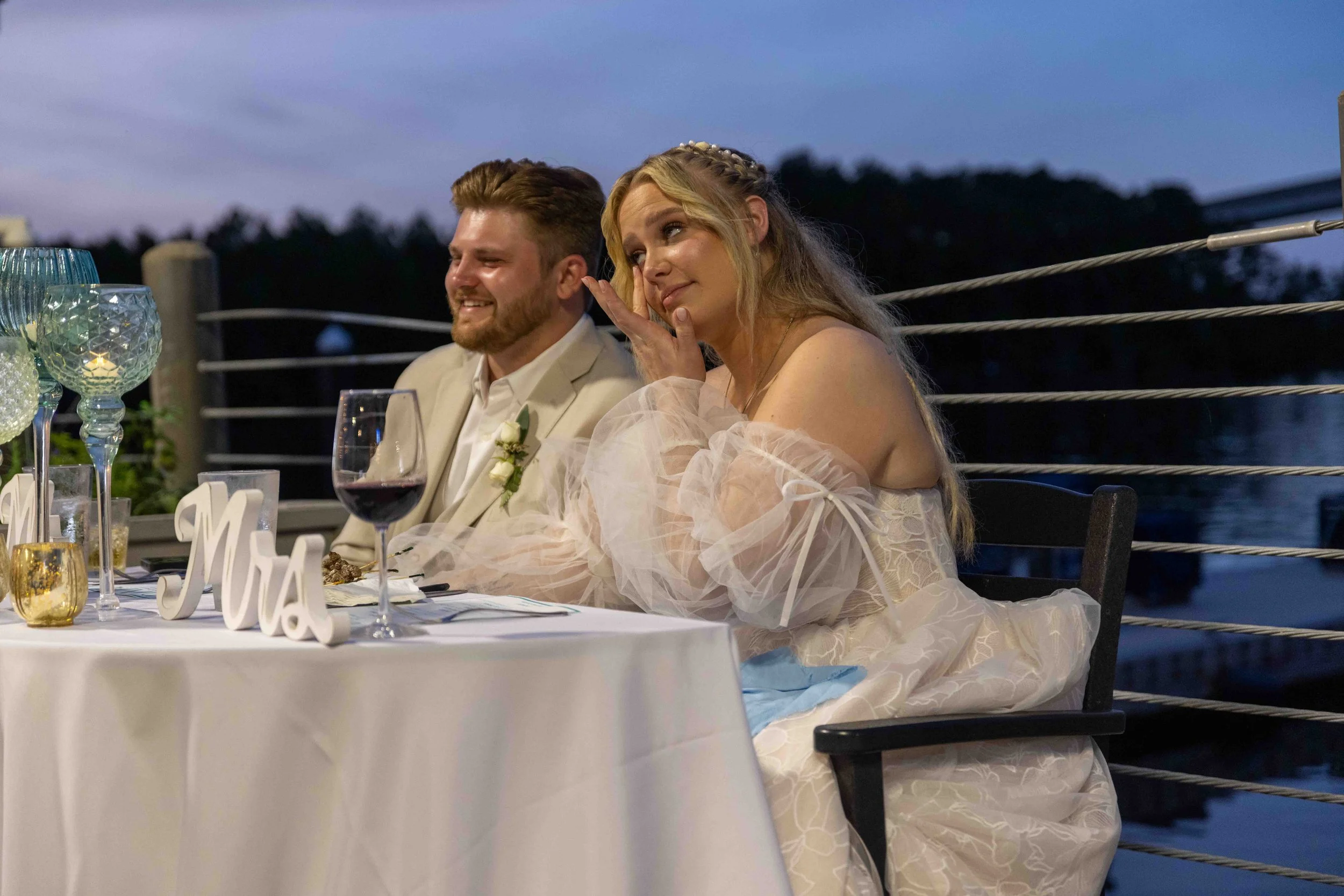 A bride and groom sitting at a wedding reception table outdoors during dusk. The bride has long platinum blonde hair styled with braids and is wearing a light-colored dress with puffy sleeves. The groom has short brown hair and a beard, and is dresse