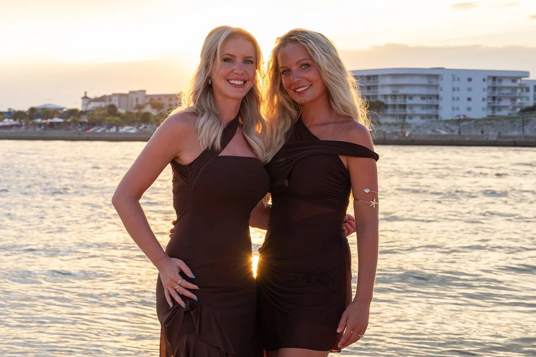 Two women in black dresses smiling by the water at sunset with buildings in the background.
