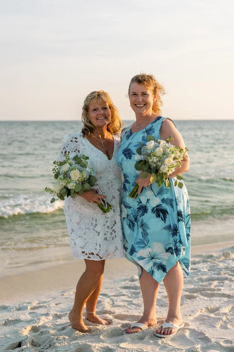 Two women standing on the beach in front of the ocean, holding bouquets of flowers, smiling, during sunset or late afternoon.