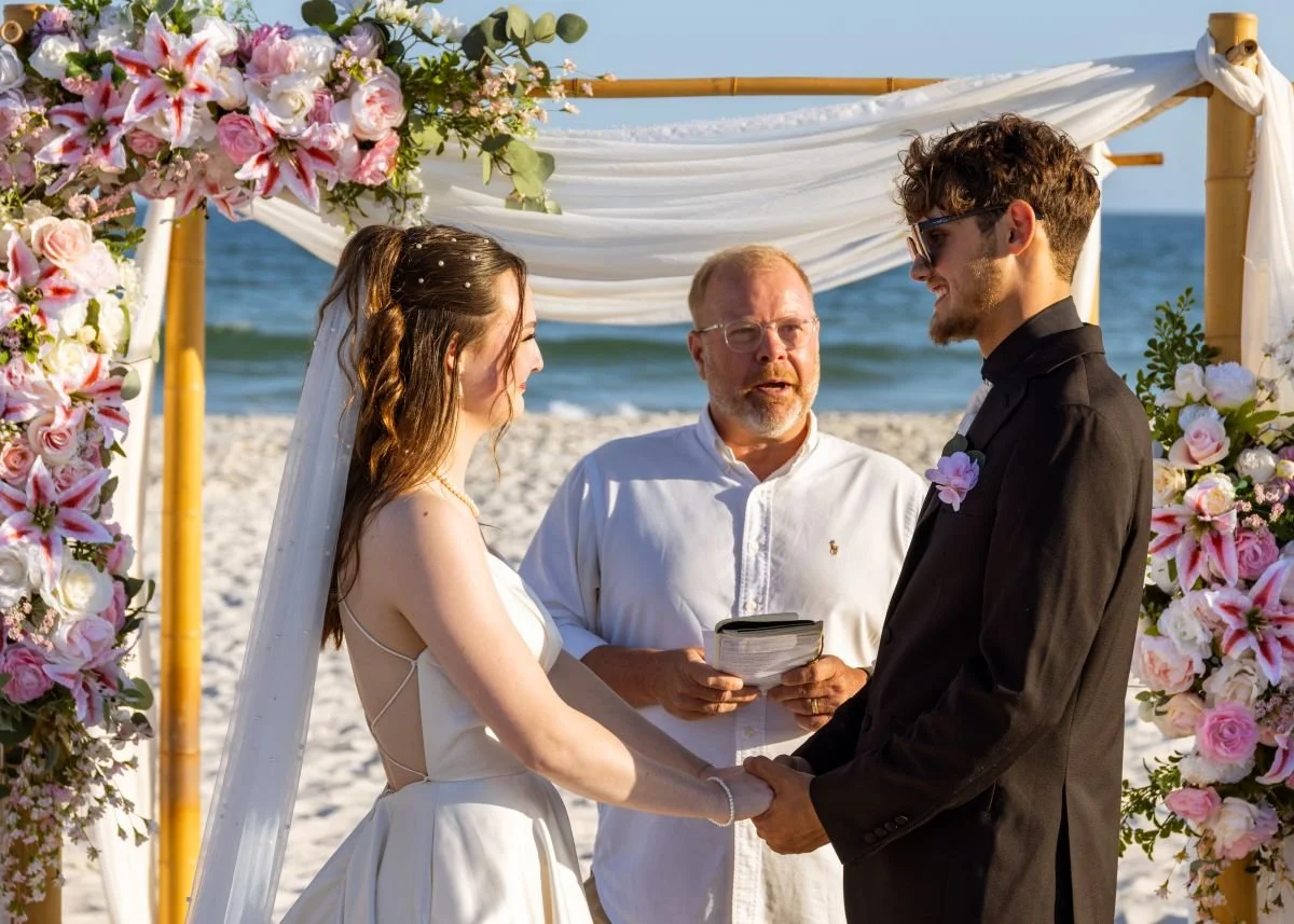 A couple gets married on the beach with an officiant during a sunny day. The bride has long brown hair and is wearing a white wedding dress with a veil, holding hands with the groom who has curly brown hair, sunglasses, and a black suit. They stand u