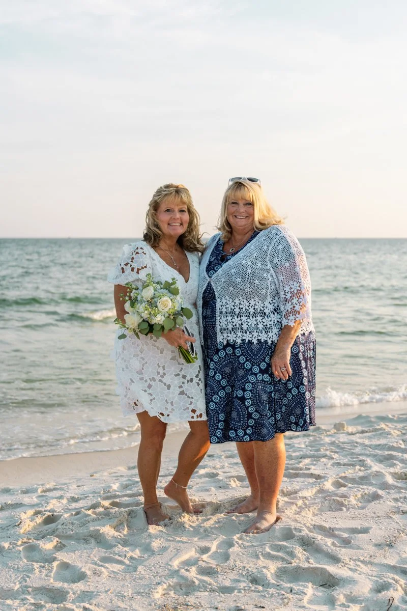 Two women standing barefoot on the beach near the ocean, smiling at the camera, one holding a flower bouquet, the other wearing sunglasses and a blue patterned dress with a white lace cover-up.